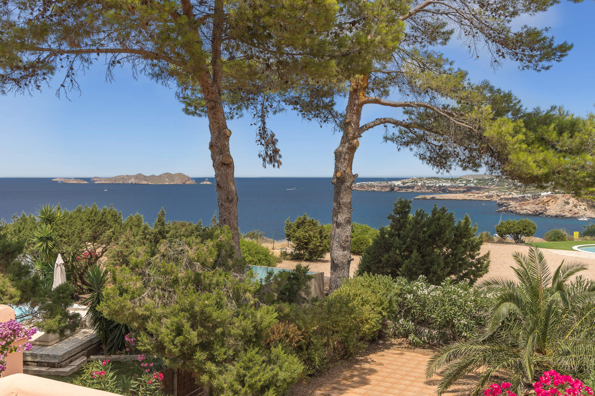 View of the blue Mediterranean sea and distant islands framed by pine trees and lush garden greenery on a sunny day.