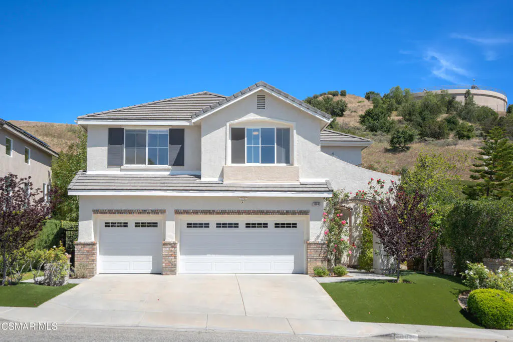 Two-story house with a two-car garage, white exterior, gray roof, and green lawn under a clear blue sky.
