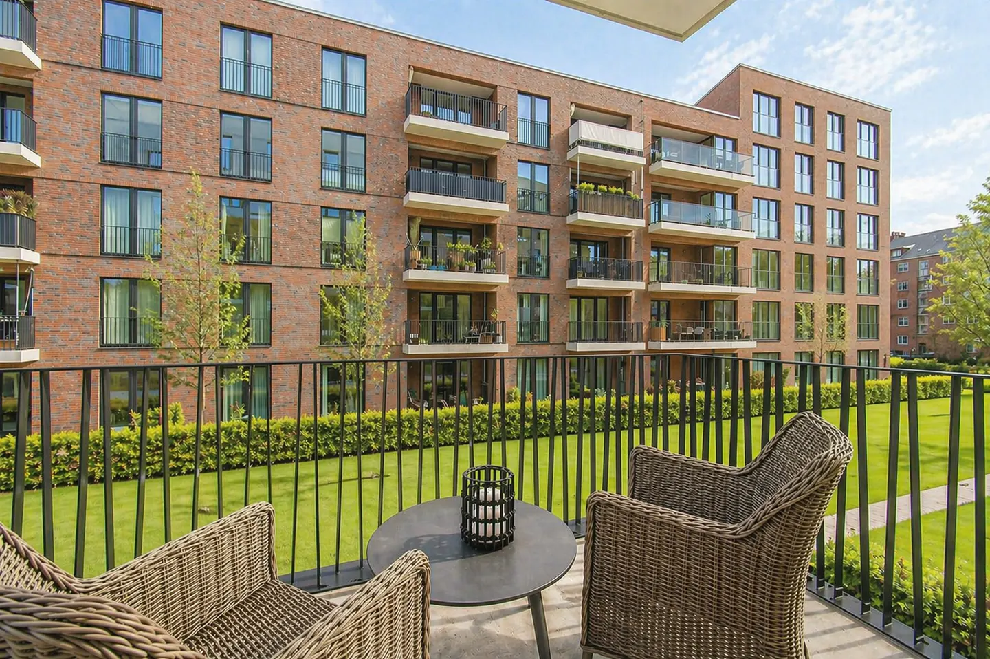 Balcony view of a brick apartment building with wicker chairs and a table overlooking a green lawn.