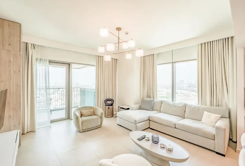 Bright, modern living room with a cream sectional sofa, matching armchair, oval coffee table, and large windows with drapes.