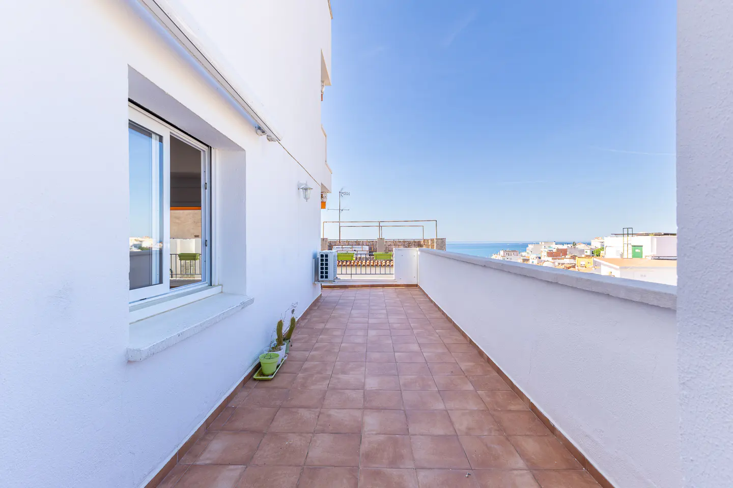 A bright, tiled balcony with white walls overlooks a coastal town and the sea under a clear blue sky.