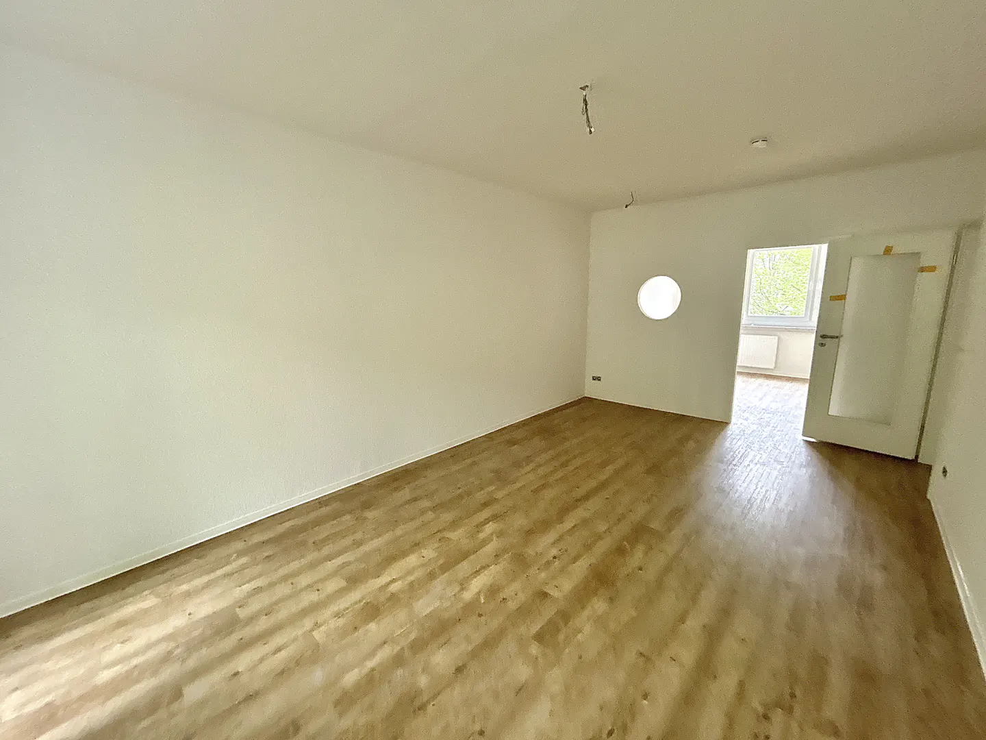 Empty room with light wood floors, white walls, and a round window. A door is ajar, revealing a glimpse of another room.