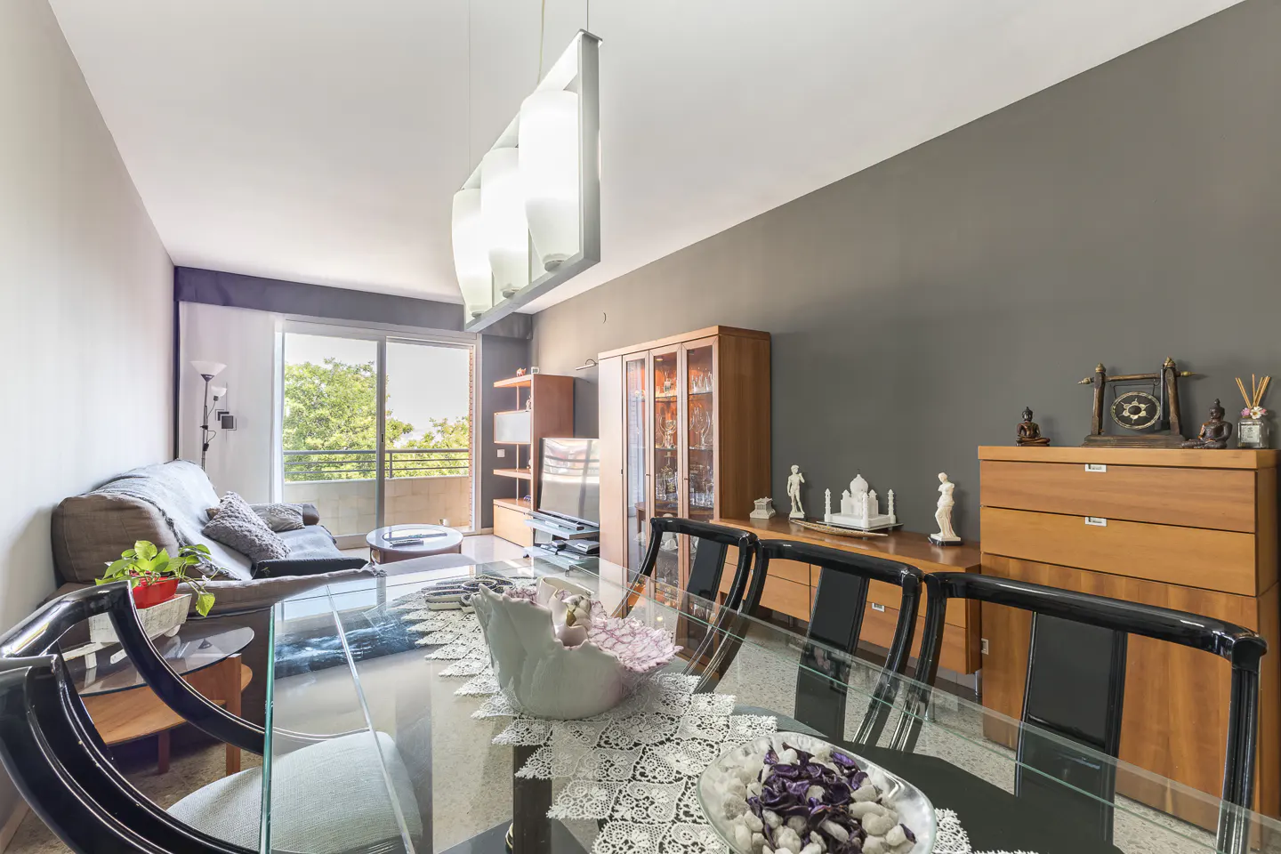 Living room with a glass dining table, black chairs, and a gray sofa near a balcony with green trees outside.