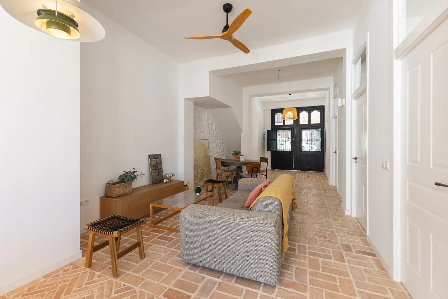 Bright living room with brick floors, white walls, and a gray sofa. A wooden ceiling fan and modern lighting fixtures add warmth to the space.