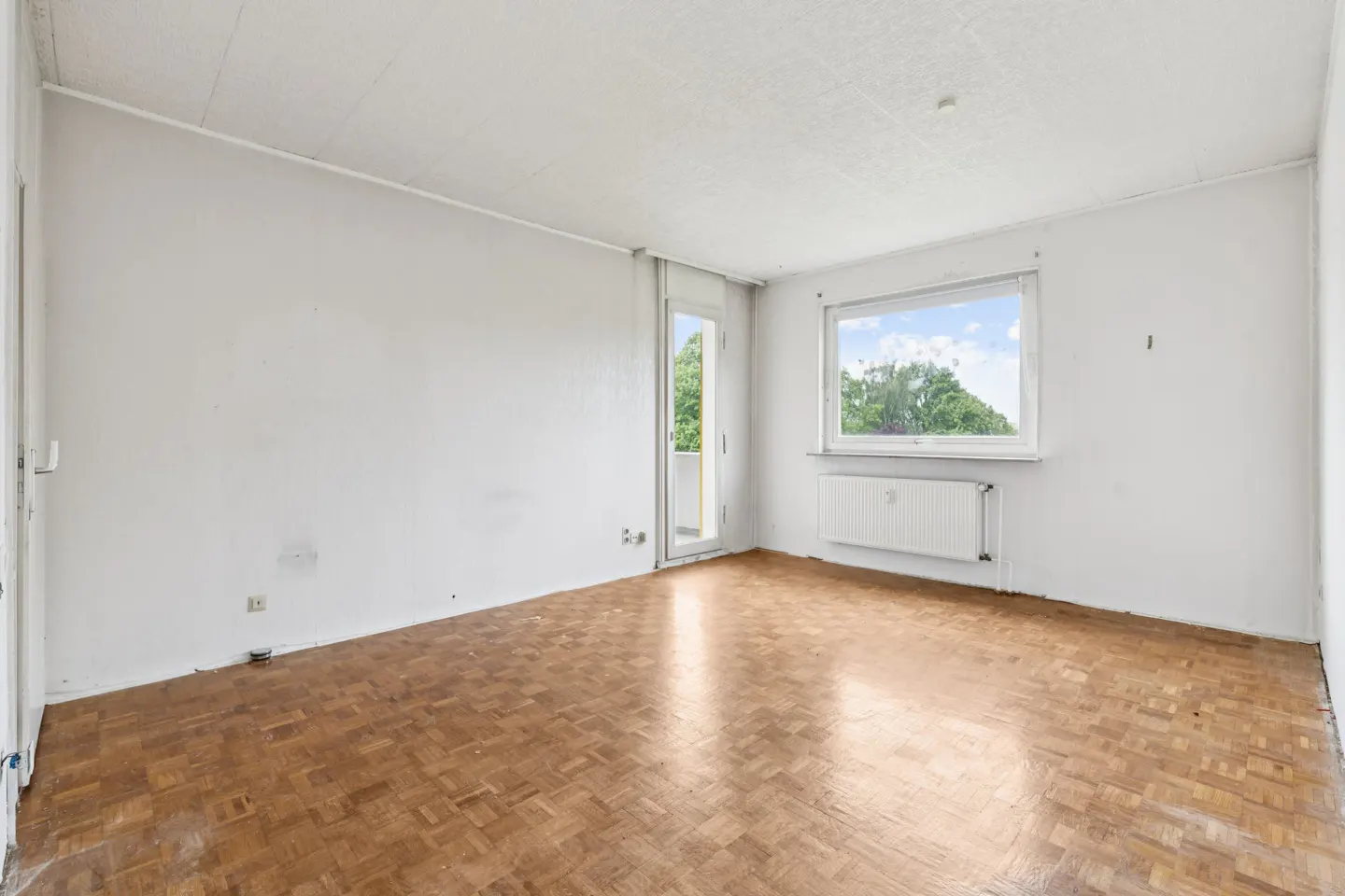 Bright, empty room with parquet floors, white walls, and a window showing trees. A door leads to a balcony.