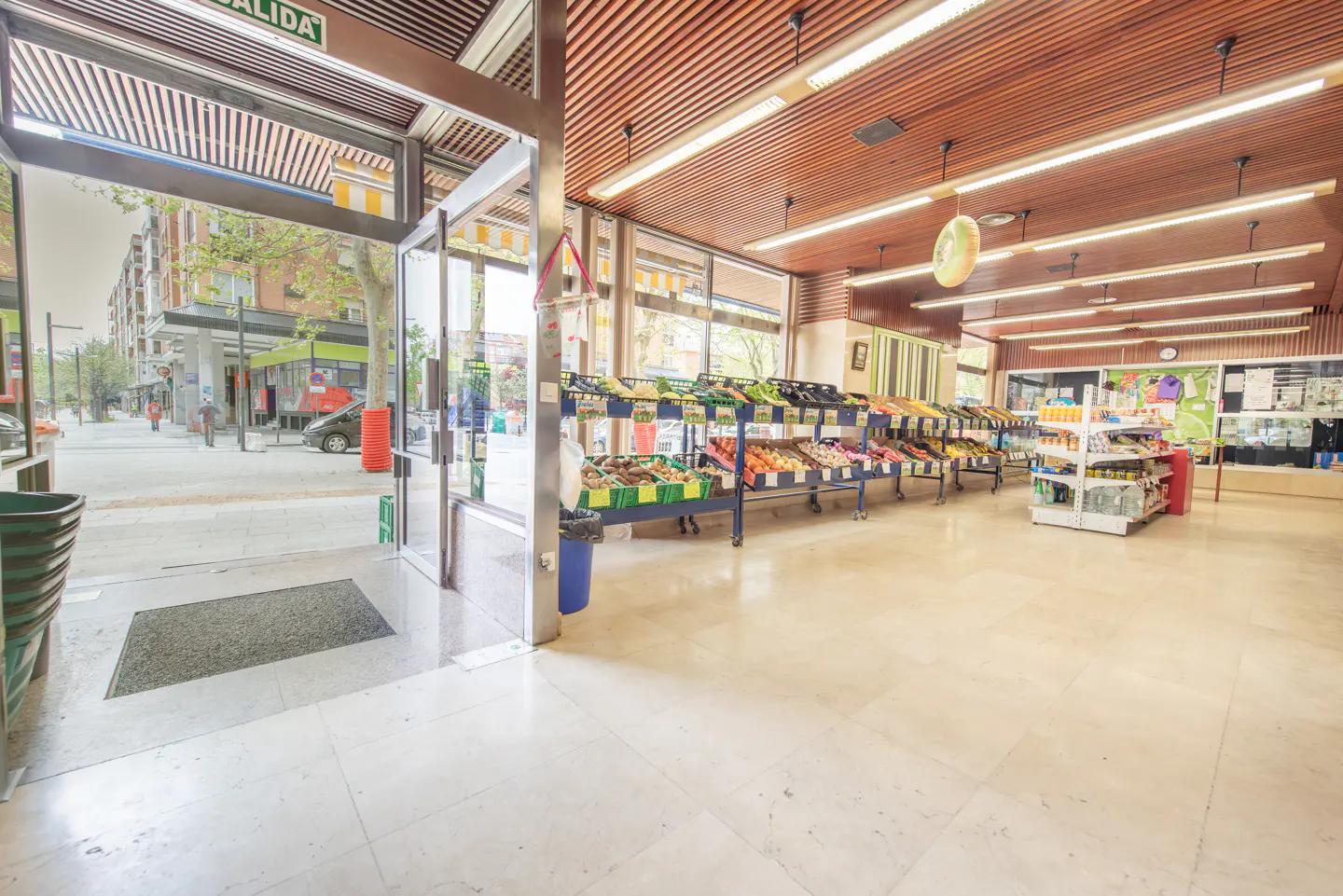 Interior view of a bright grocery store with fruit and vegetable displays. Glass doors open to a street scene. The ceiling is wood with fluorescent lighting.