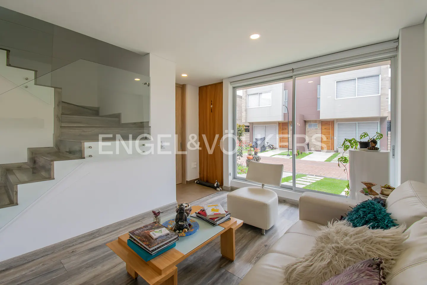 Bright living room with white sofa, wood floors, and glass-railed stairs. Large window overlooks a courtyard with townhouses.