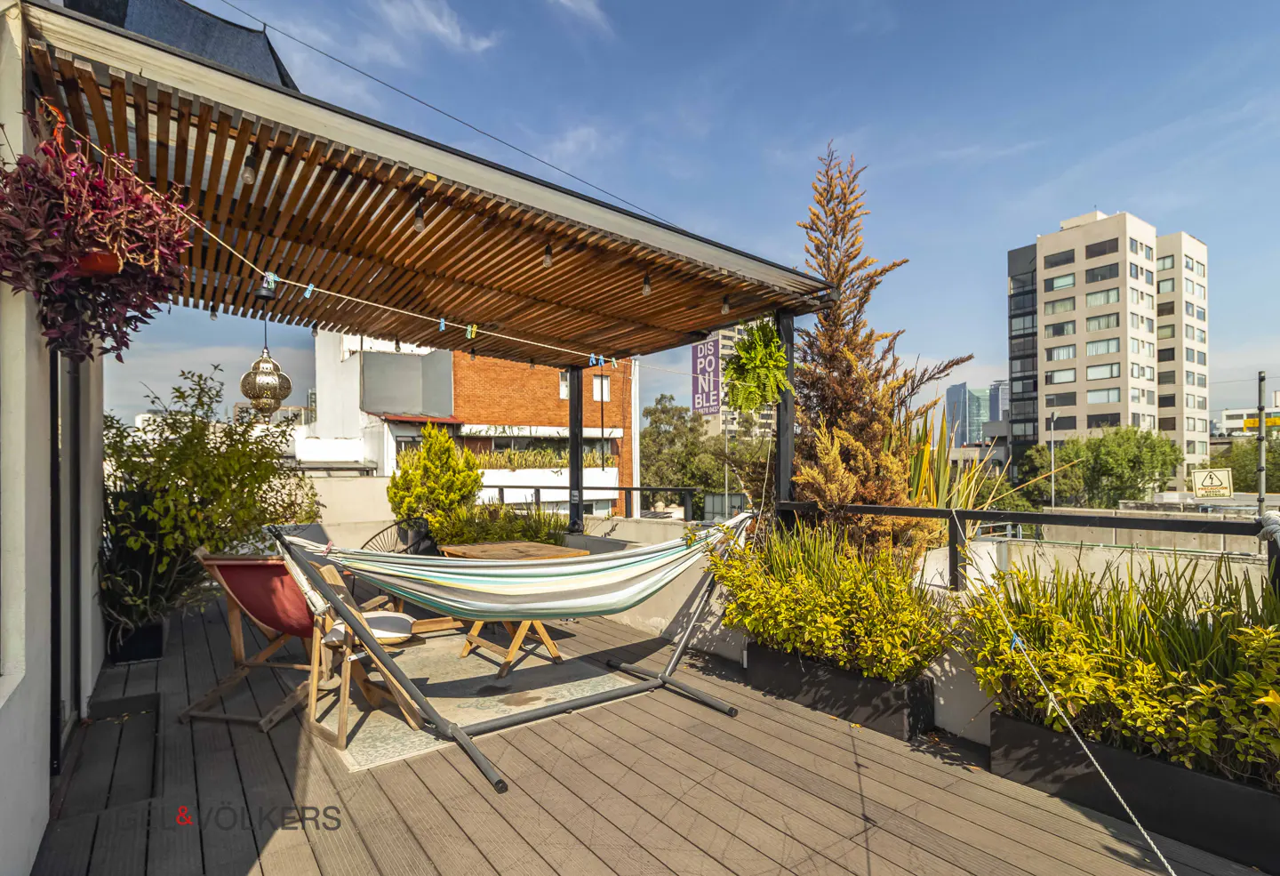 Rooftop patio with a hammock, chairs, and plants under a wooden pergola. City buildings are visible in the background.