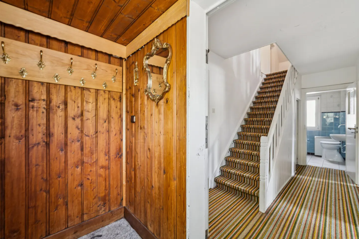 A home's interior shows a wood-paneled entryway with gold hooks and a mirror, leading to a carpeted staircase and a glimpse of a blue-tiled bathroom.