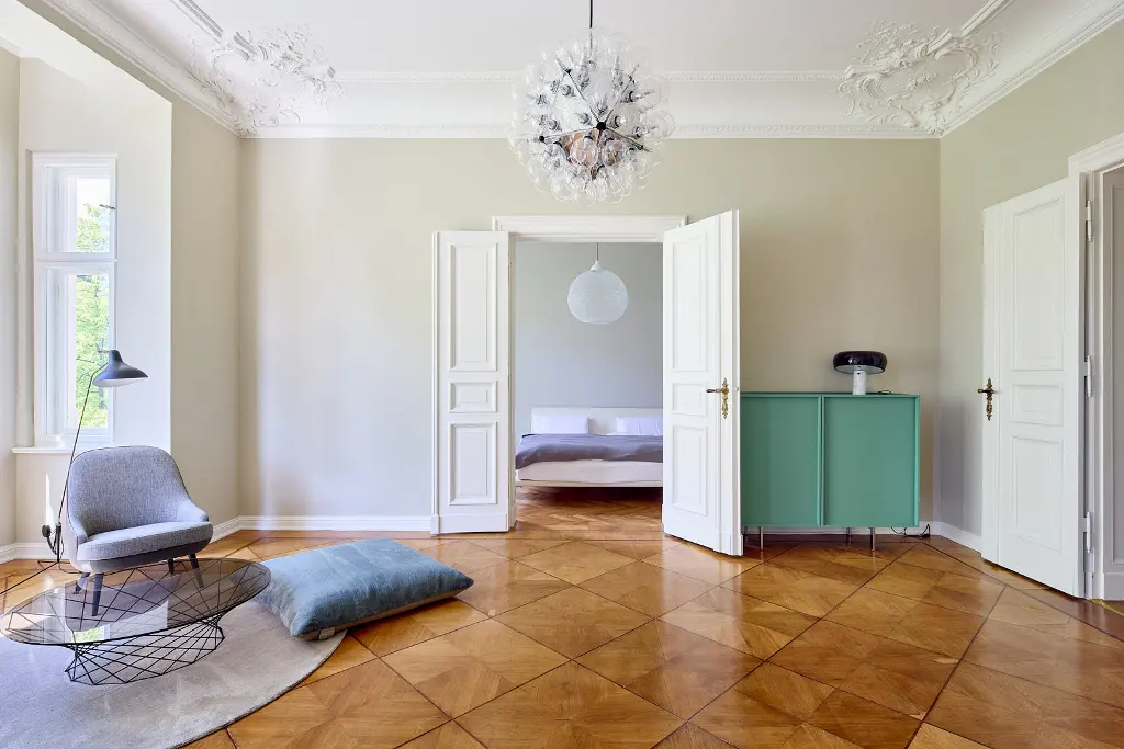 Bright living room with parquet floors, a gray chair, and a blue cushion. Open doors lead to a bedroom with a white bed. A green cabinet stands against the wall.
