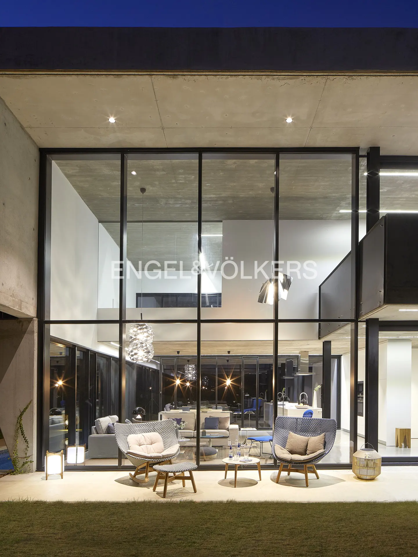 Modern home exterior at dusk with large glass windows, patio furniture, and Engel & Völkers logo visible inside.