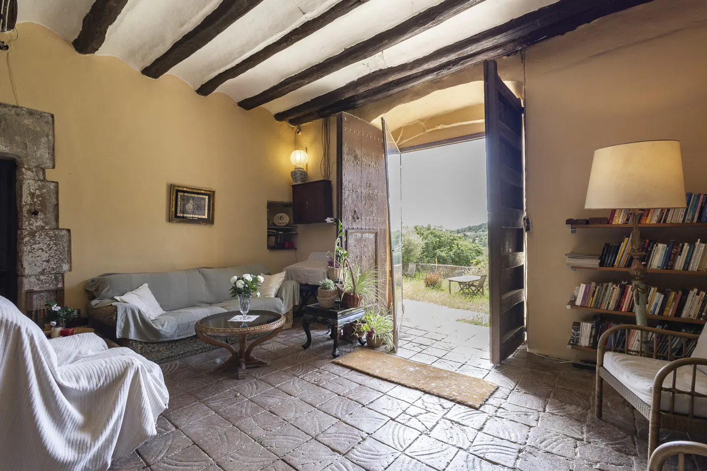 Living room with exposed beams, stone floors, and open doorway to a garden. A sofa, table, and bookshelf furnish the room.
