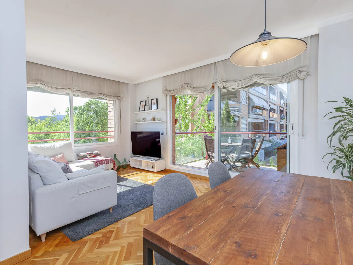 Bright living room with a white sofa, TV, wood table, and balcony with chairs. A large window shows green trees outside.
