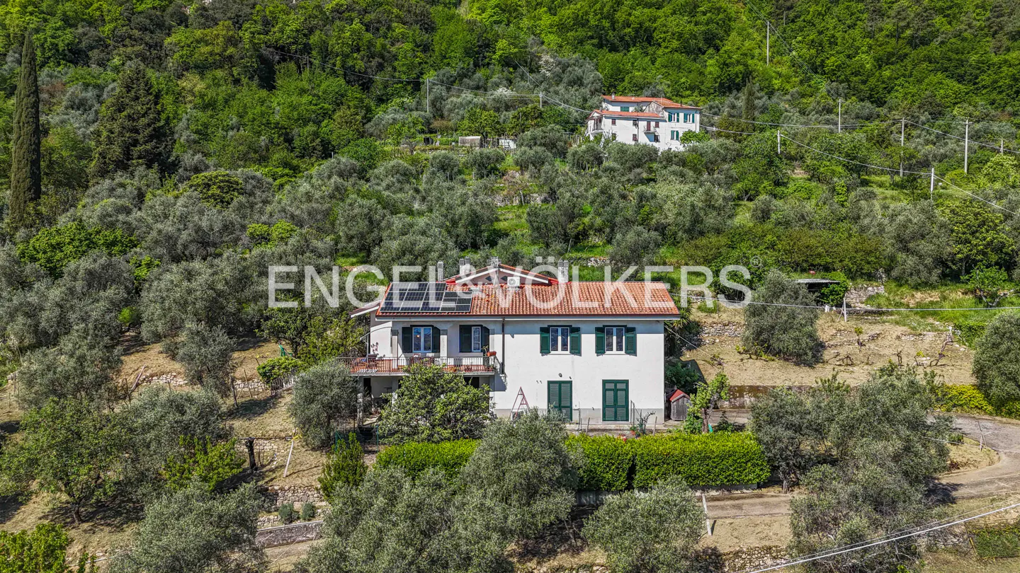 A white two-story house with green shutters and a red tile roof sits on a green hillside.