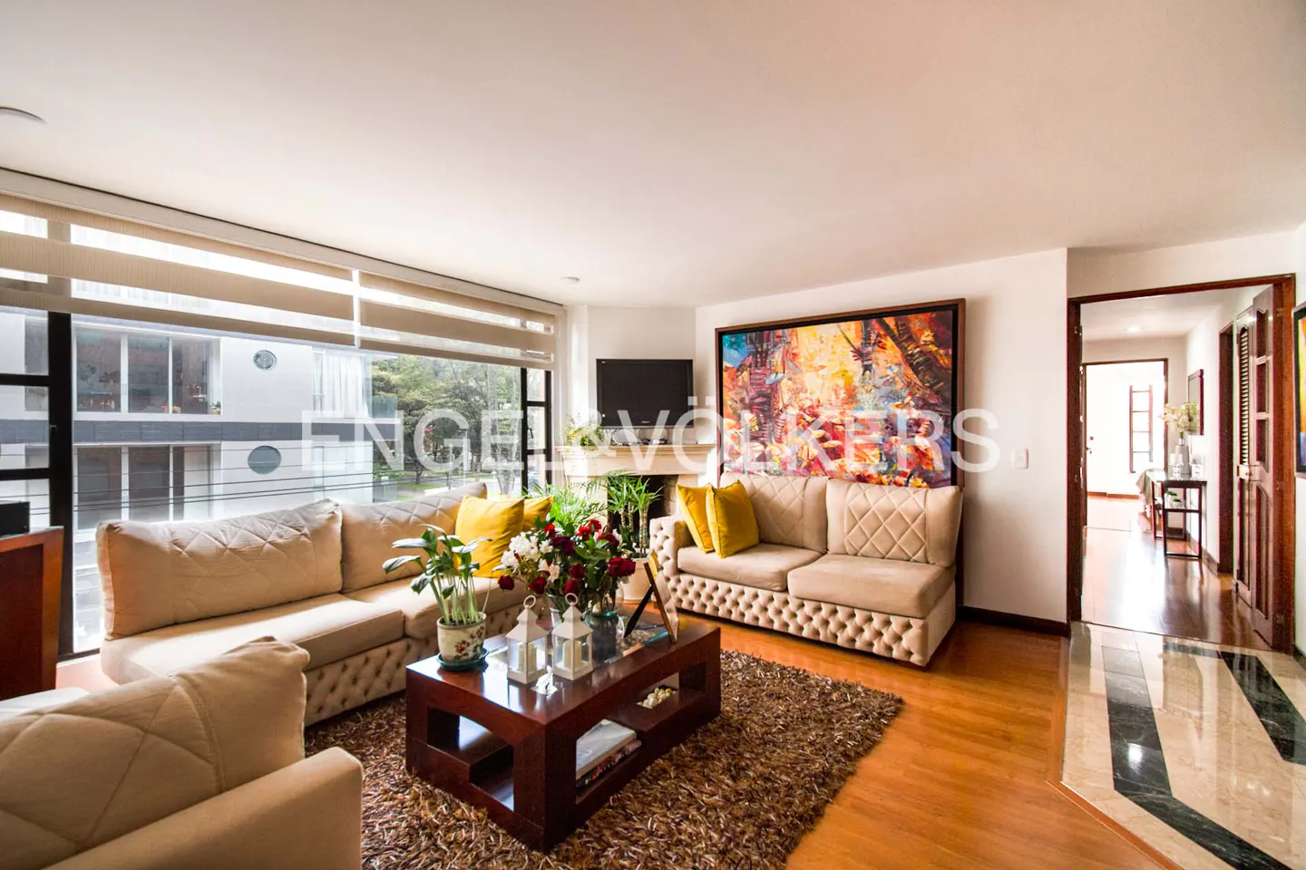 Bright living room with tan sofas, yellow pillows, and a dark wood coffee table on a brown rug. A colorful painting hangs above the sofa.