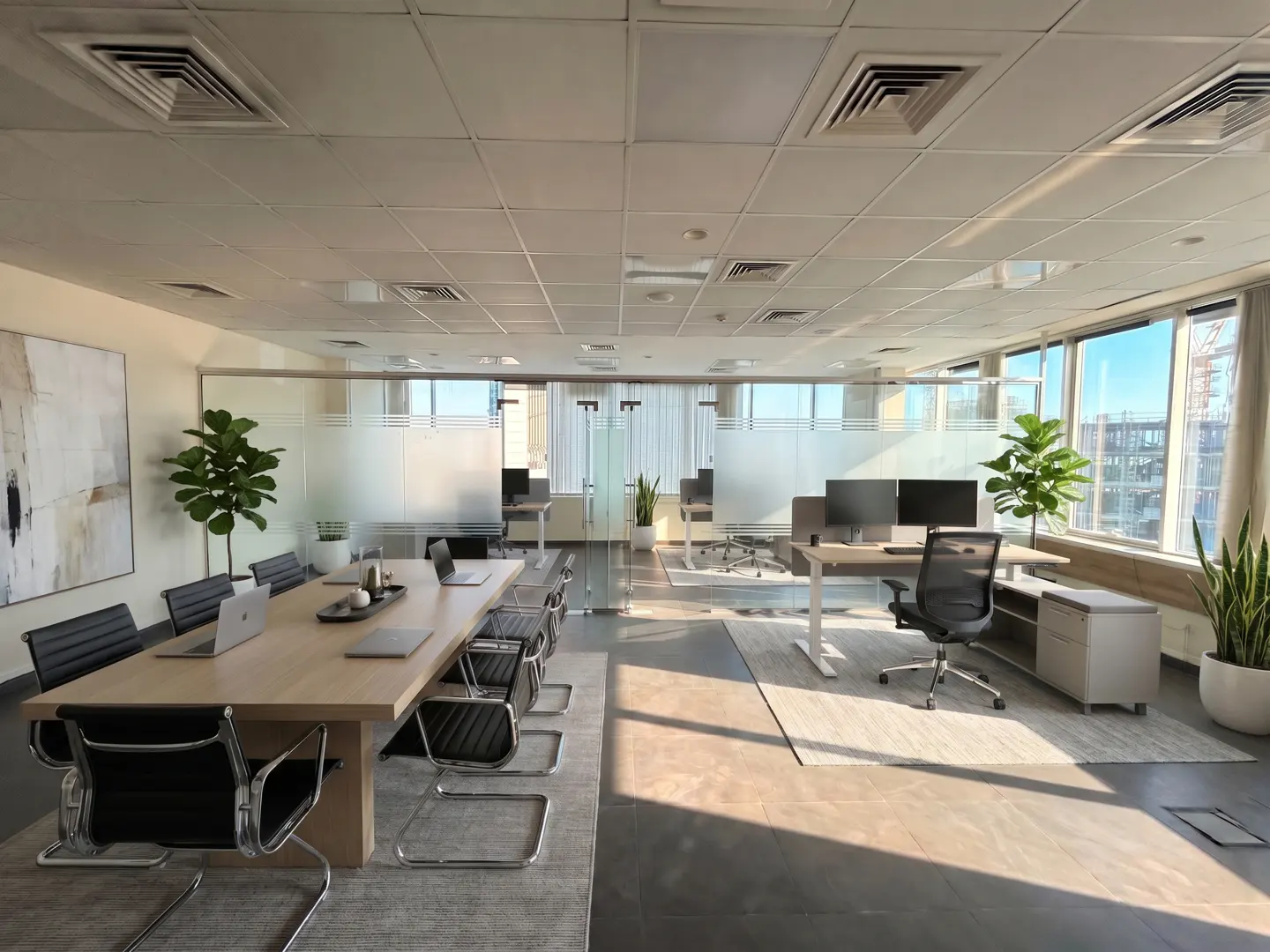 Bright, modern office space with a long conference table, black chairs, and individual workstations with computers. Large windows offer natural light.
