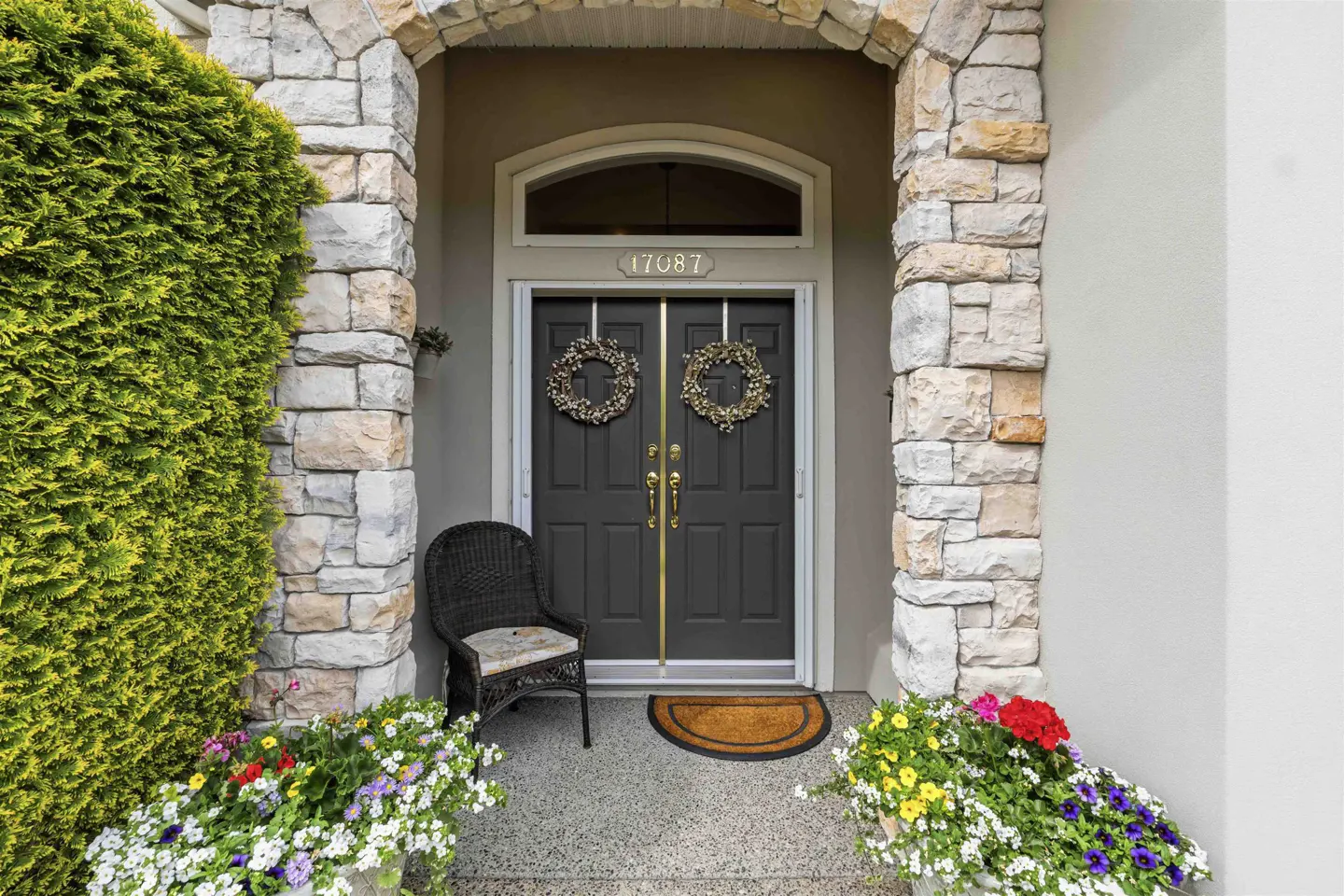 Front entrance of a house with gray double doors, stone archway, wreaths, and colorful flowers.