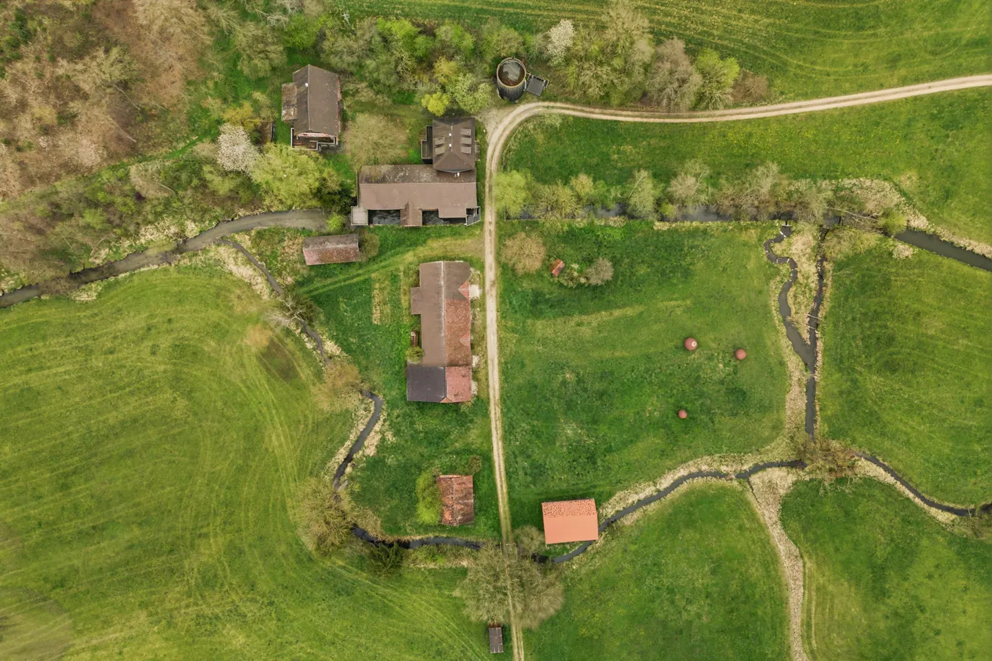 Aerial view of a rural property with several brown buildings, green fields, a dirt road, and a small stream.