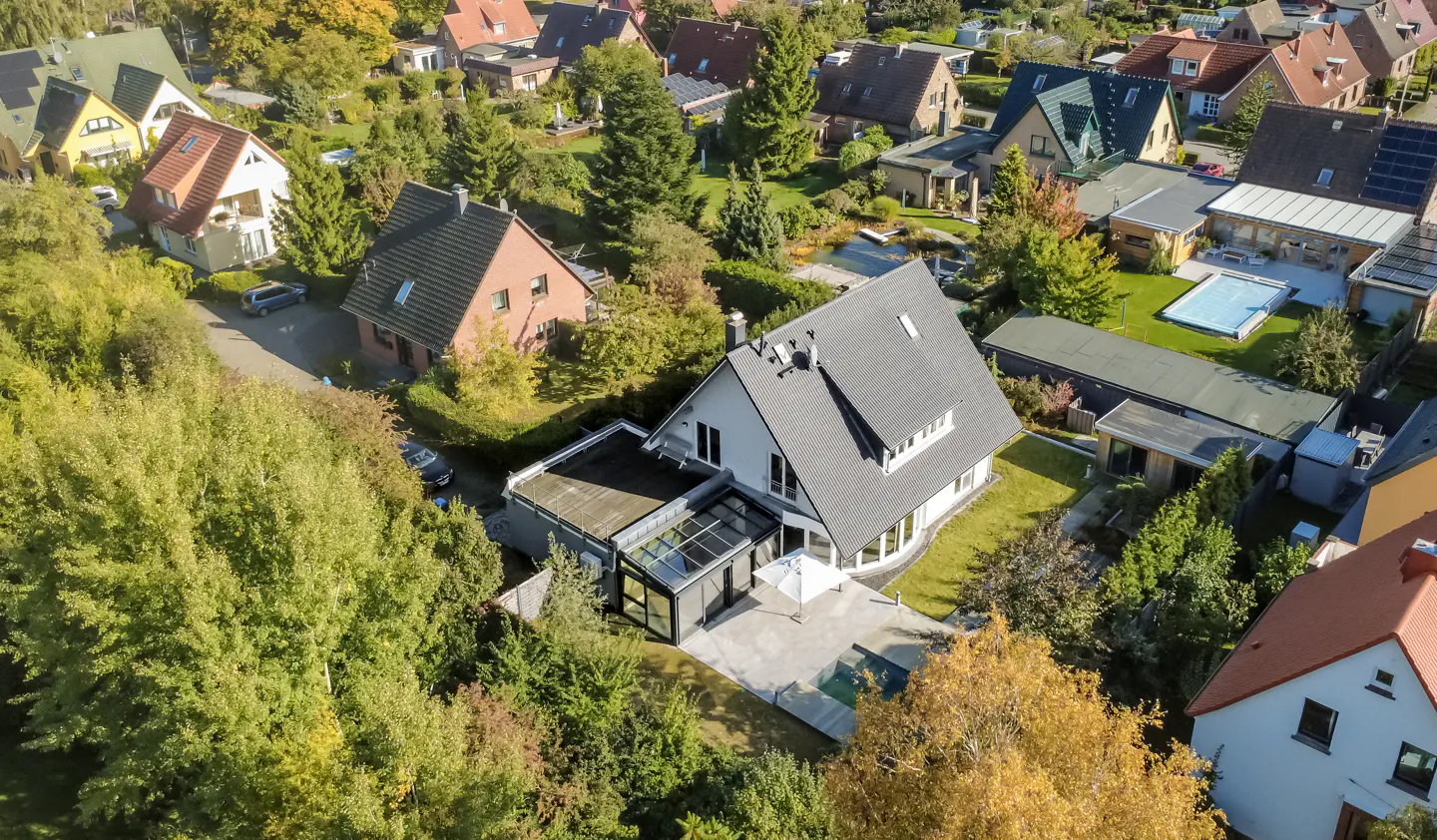 Aerial view of a modern white house with a gray roof, surrounded by green trees and other houses in a neighborhood.