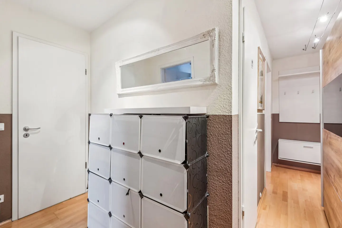 Hallway with a white door, a modular shoe cabinet, and a framed mirror on a two-tone wall. A glimpse into another room with wooden floors.