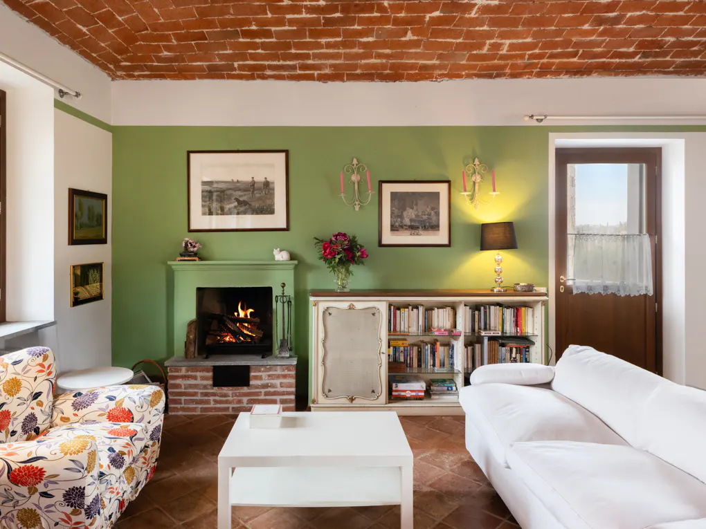Living room with brick ceiling, green walls, and terracotta tile floor. A fireplace, bookshelves, and white sofa furnish the space.