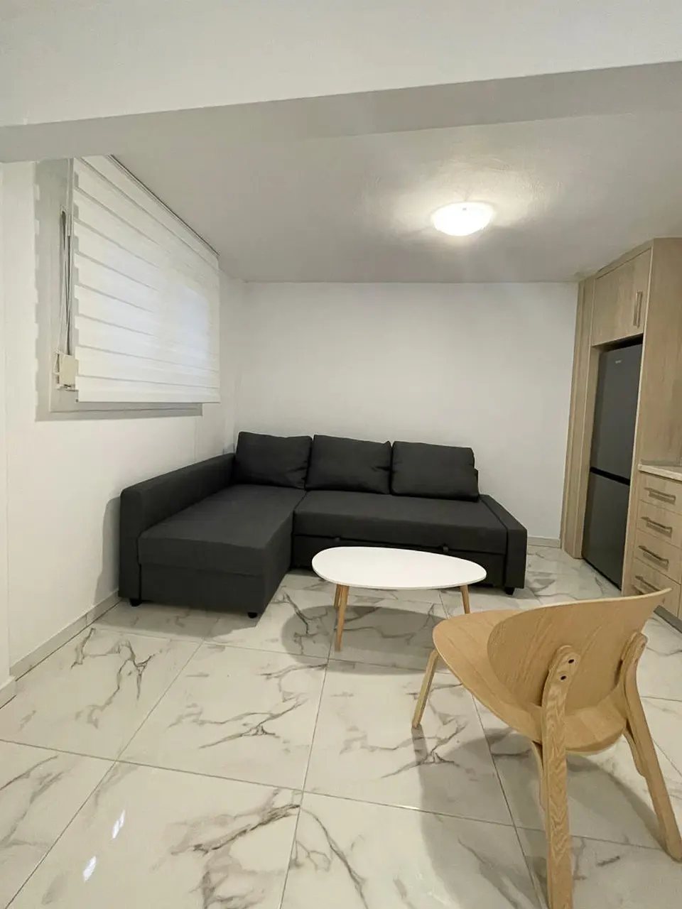 Living room with a gray sectional sofa, white coffee table, and wooden chair on marble tile floor. Kitchen cabinets in the background.