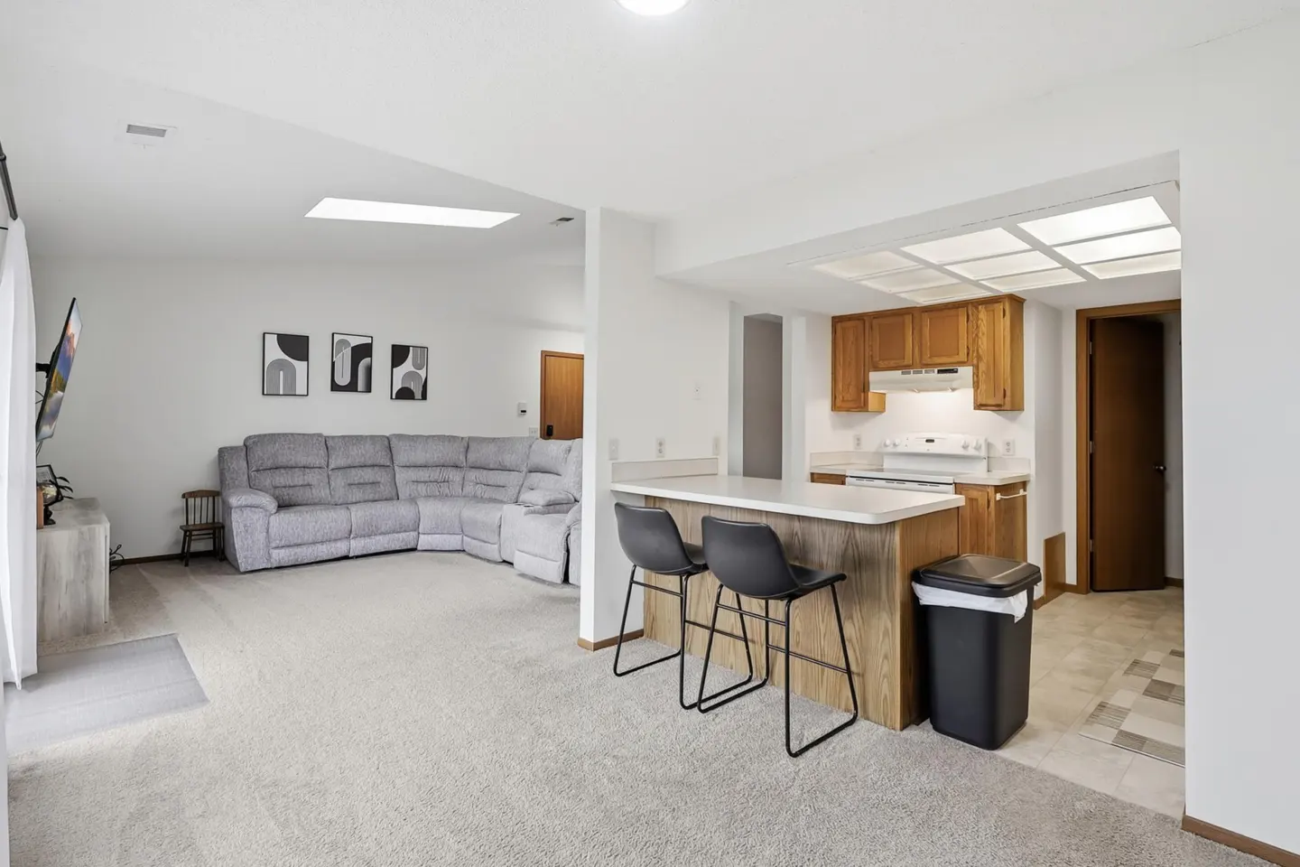 Open-concept living space with a gray sectional sofa, kitchen bar with two black stools, and oak cabinets.
