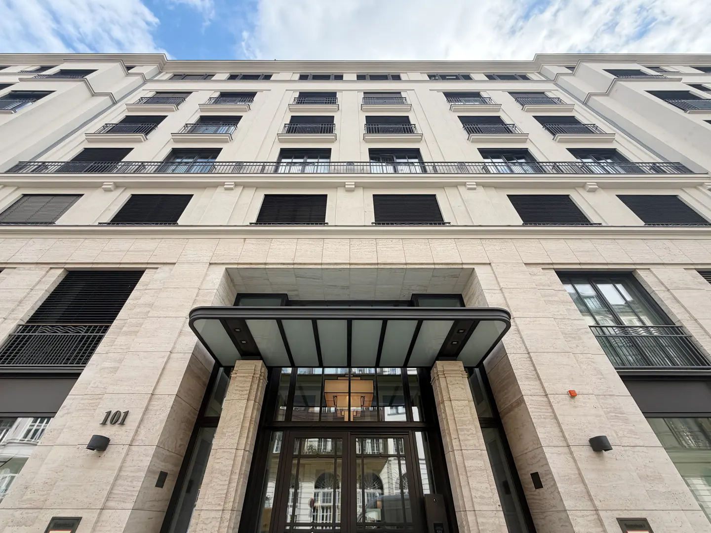 Low angle view of a tall, light-beige building with black window shutters and balconies. The entrance has a glass awning and the number 101 is visible.