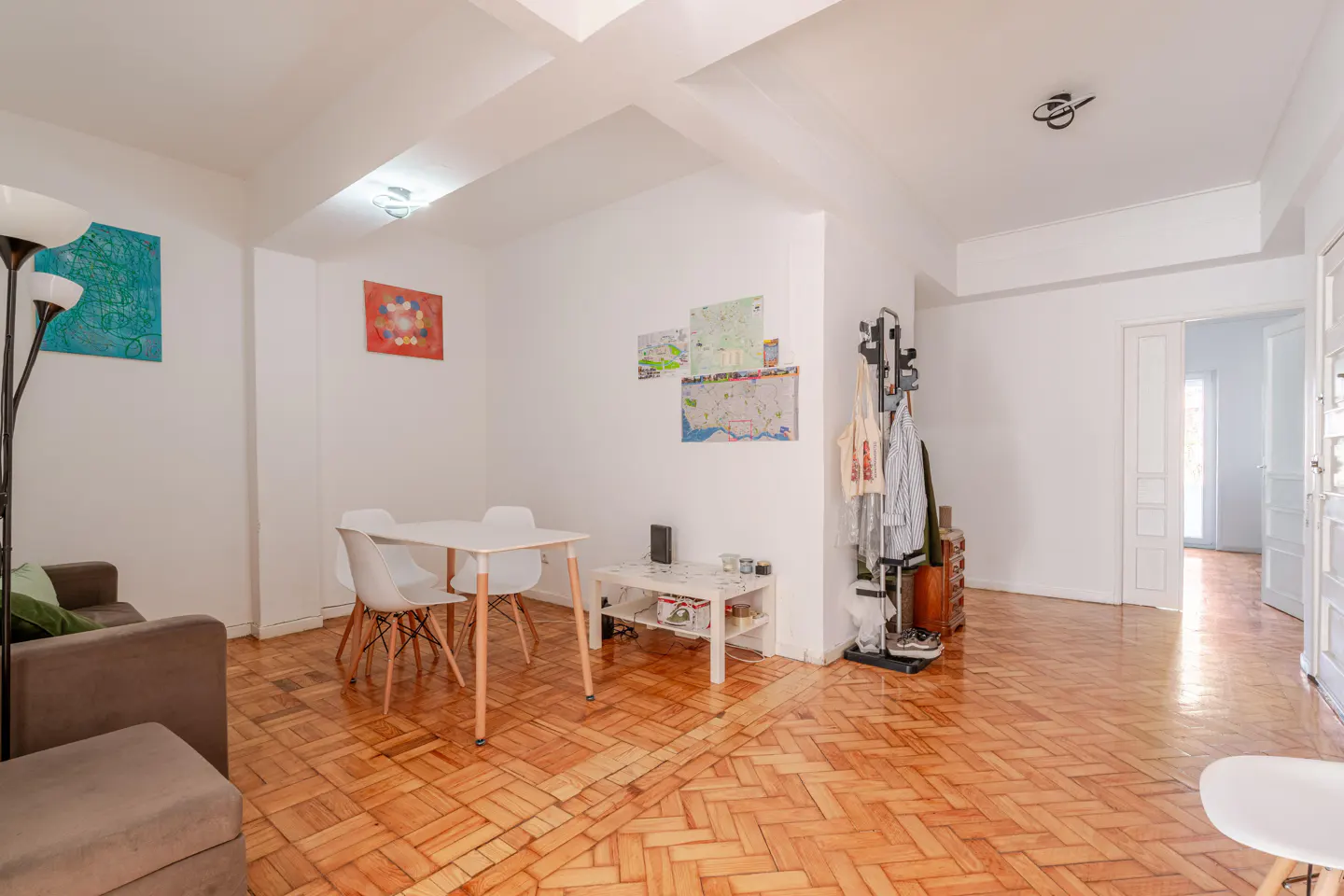 Bright living room with parquet floors, white walls, and modern furniture. A gray sofa and white dining set are visible. Maps and art decorate the walls.