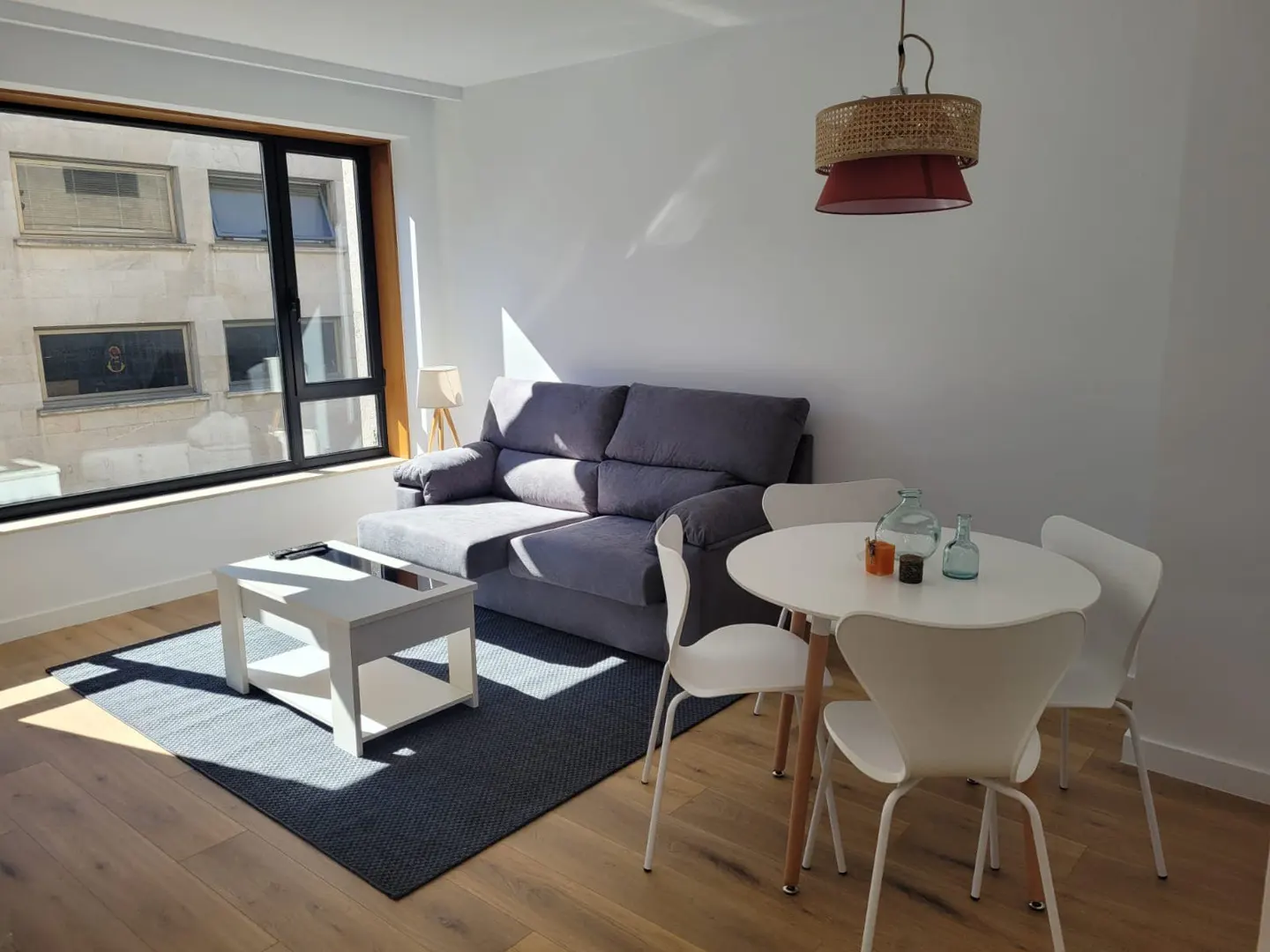 Bright living room with a gray sofa, white table and chairs, and a large window with sunlight streaming in.