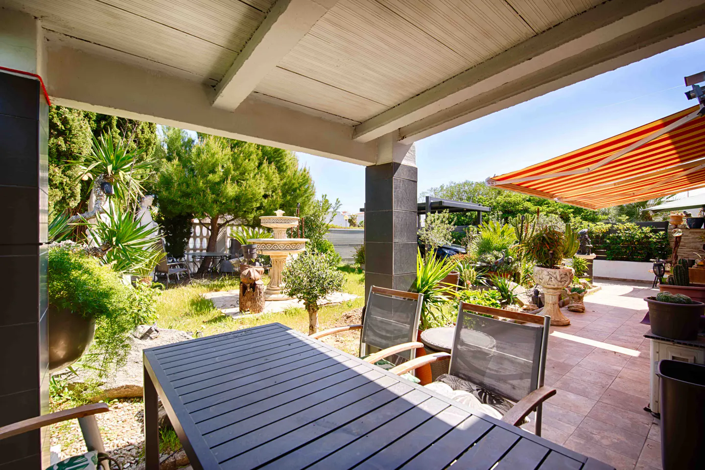 Covered patio with a dark table and chairs overlooks a lush green garden with a fountain and red and white striped awning.