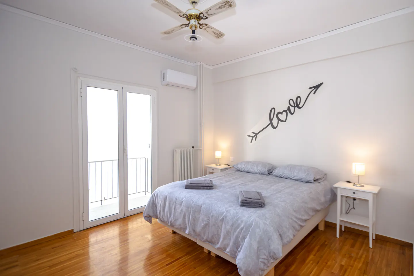 Bedroom with a gray patterned duvet, white walls, and wood floors. A "love" arrow decorates the wall. Balcony doors let in light.