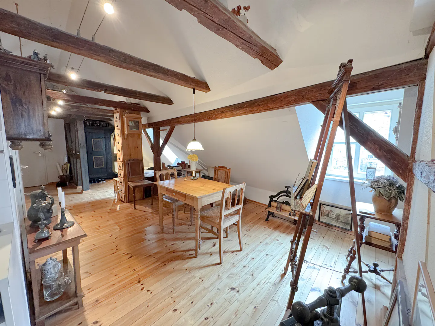 Attic apartment with exposed beams, wood floors, and a dining table with chairs. An easel stands near a window, and antique furniture adds character.