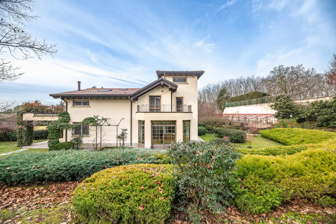 A two-story beige house with a balcony and a red tile roof, surrounded by green bushes and trees under a blue sky.