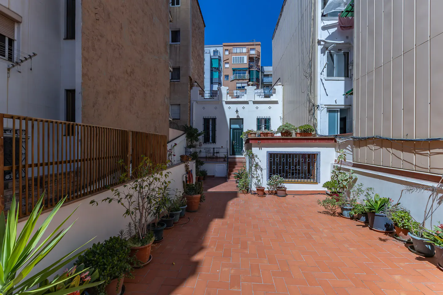 A brick-paved courtyard with potted plants, enclosed by buildings and a wooden fence. A white house with a green door is visible in the background.