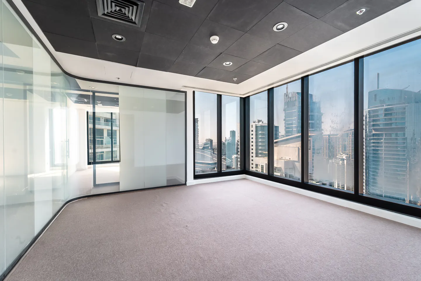 Empty office space with gray carpet, glass walls, and a black ceiling. Large windows offer a city view.