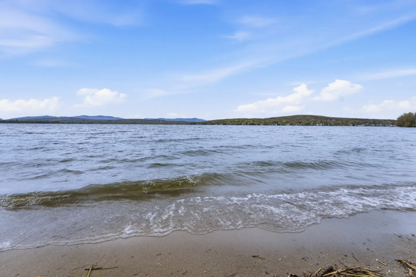 Lake view with gentle waves lapping onto a sandy shore under a blue sky with scattered clouds. Distant hills are visible.