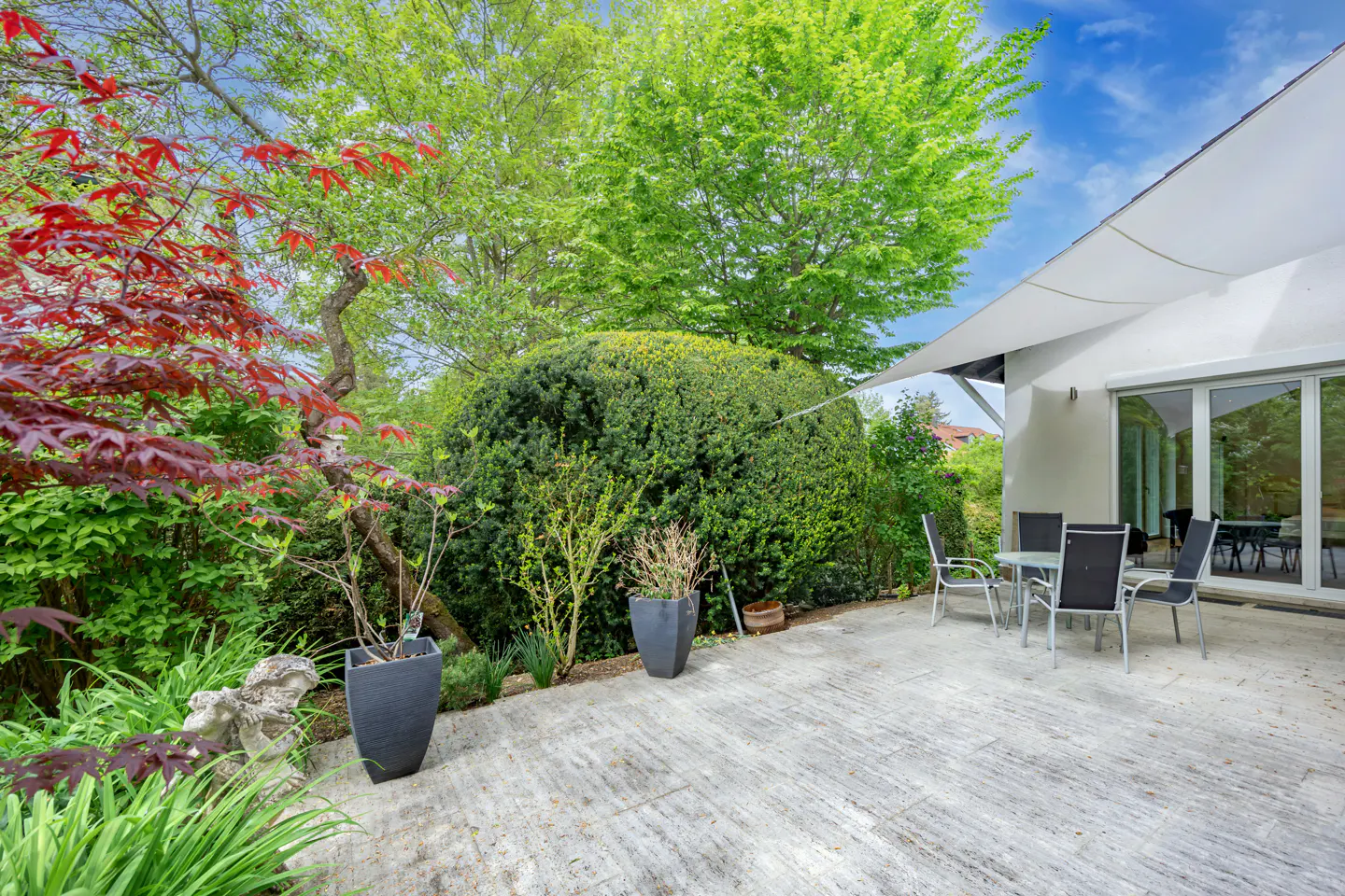A patio with a table and chairs sits outside a white house, surrounded by lush green trees and a red Japanese maple.