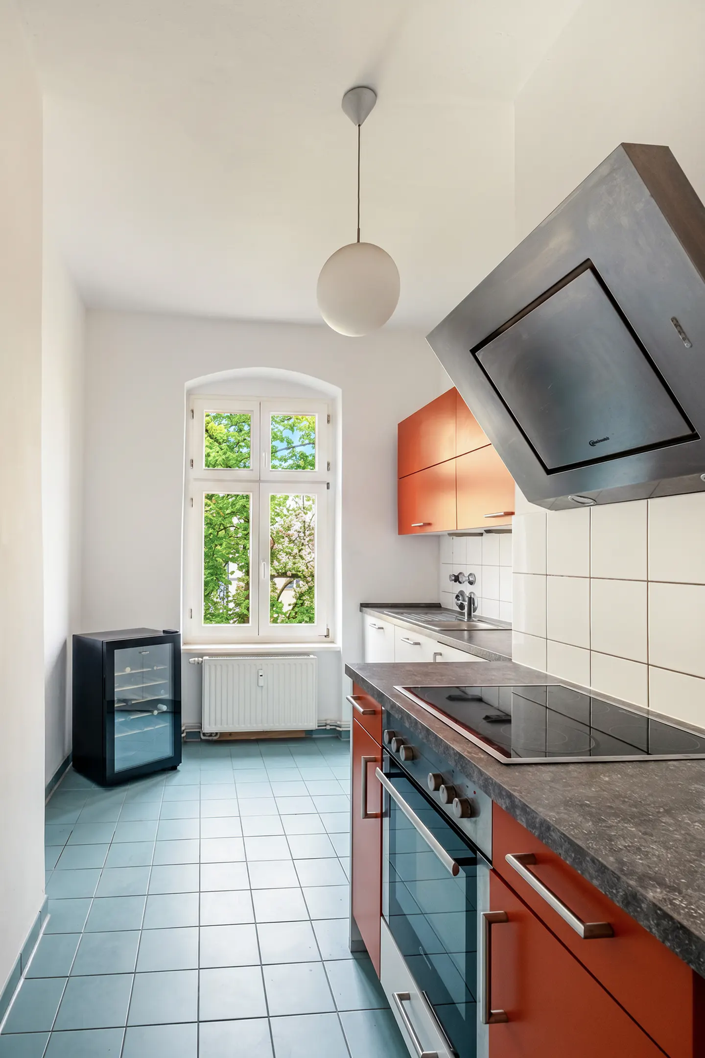 Bright kitchen with blue tile floor, white walls, and orange cabinets. A window overlooks green trees. Appliances include a black mini fridge and oven.