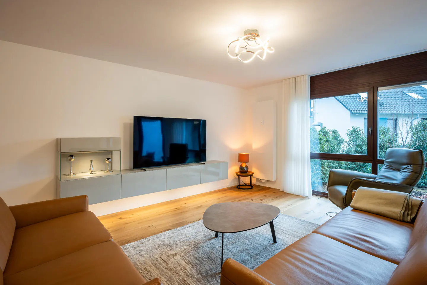 Bright living room with brown leather sofas, a gray TV stand, and a large window overlooking a green yard.