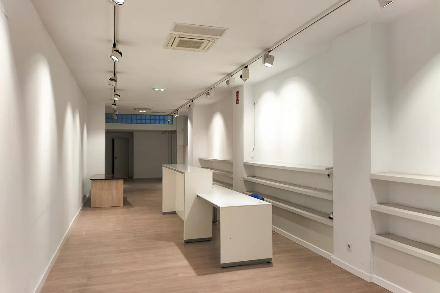 Bright, empty retail space with white walls, light wood floors, track lighting, and built-in shelving. A white counter sits in the center.