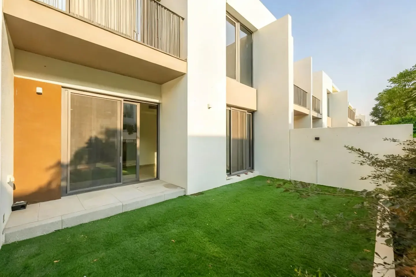 Modern townhouse exterior with a small green lawn, white walls, sliding glass doors, and a balcony above.
