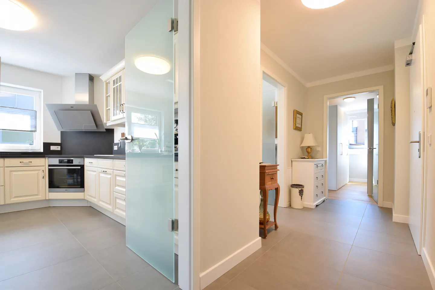 A bright, modern kitchen and hallway with gray tile floors and white walls. A frosted glass door separates the spaces.