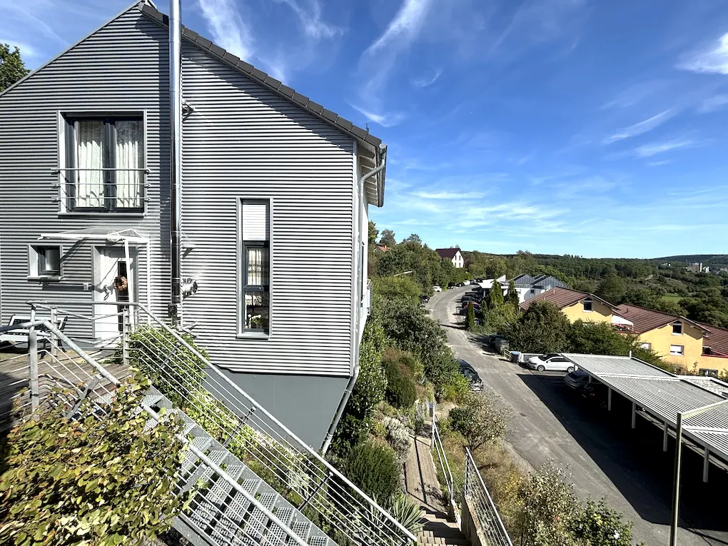 Exterior view of a modern gray house with metal siding and a metal staircase, overlooking a street with other houses and greenery.