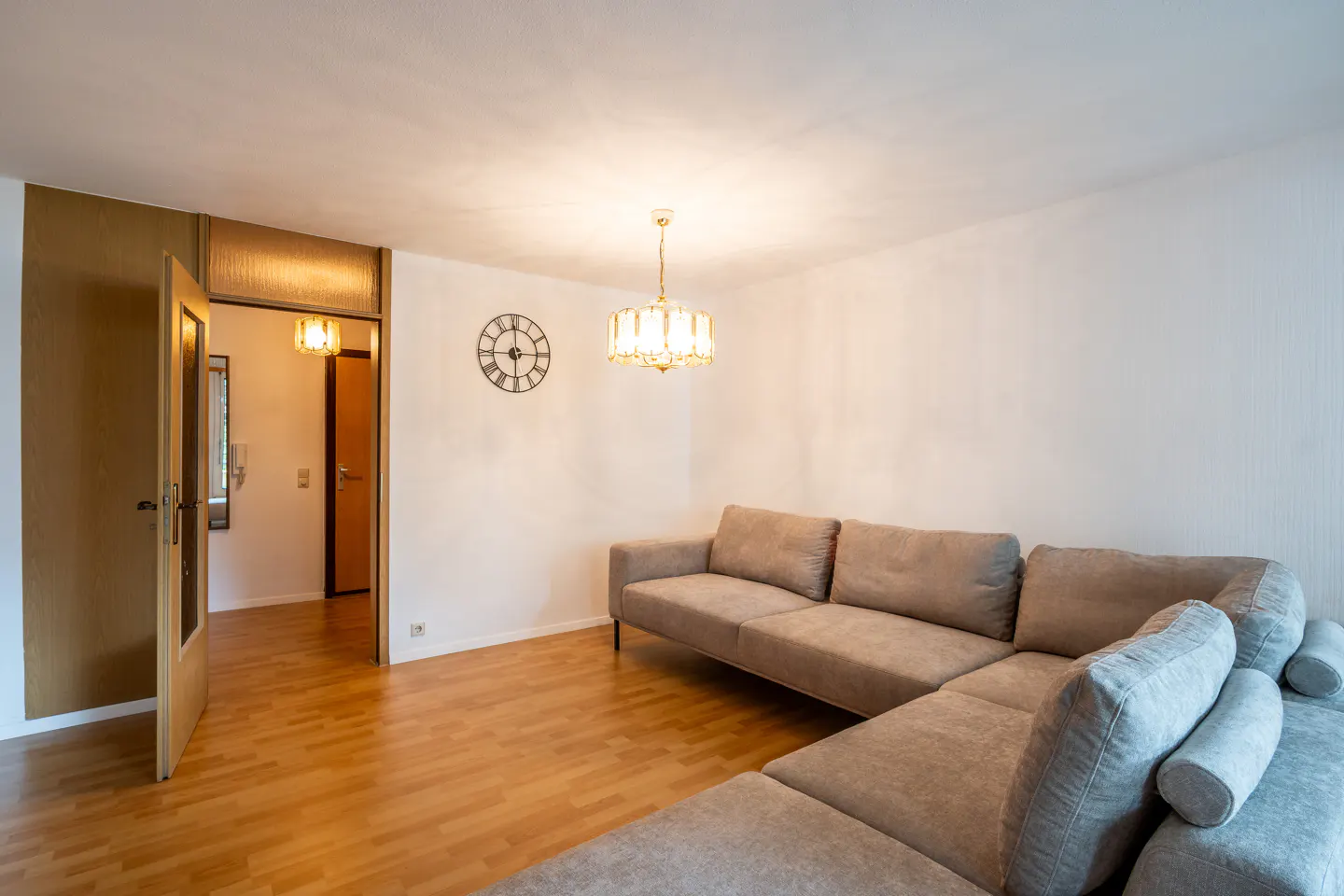 Living room with hardwood floors, a beige sectional sofa, a clock on the wall, and a chandelier.