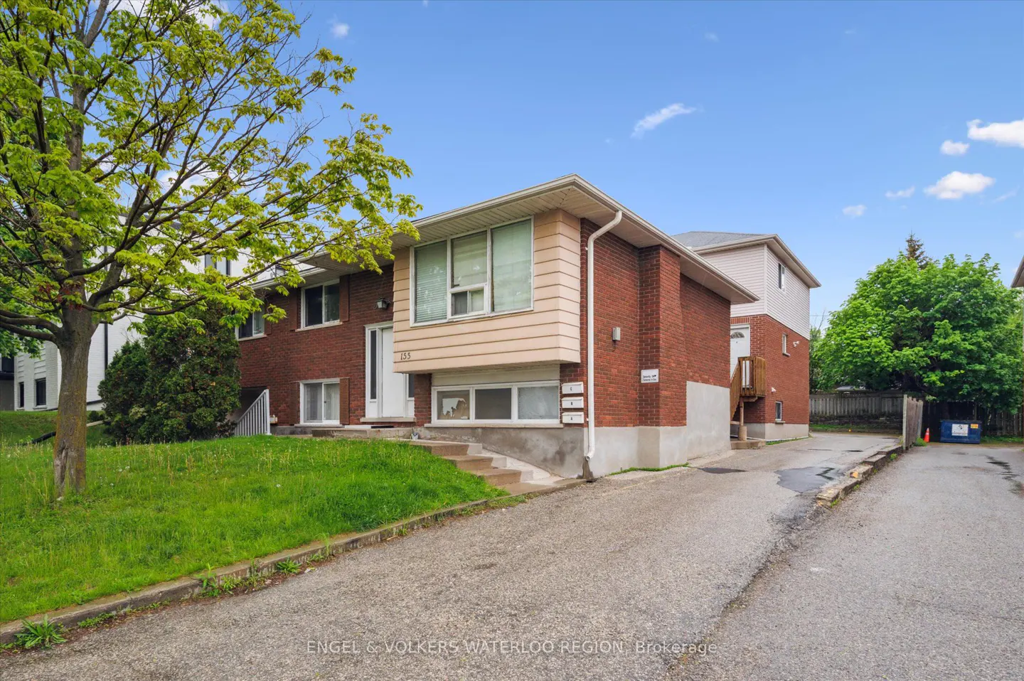 Exterior view of a two-story red brick apartment building with a beige accent. Green lawn and trees are in the front. Blue sky.