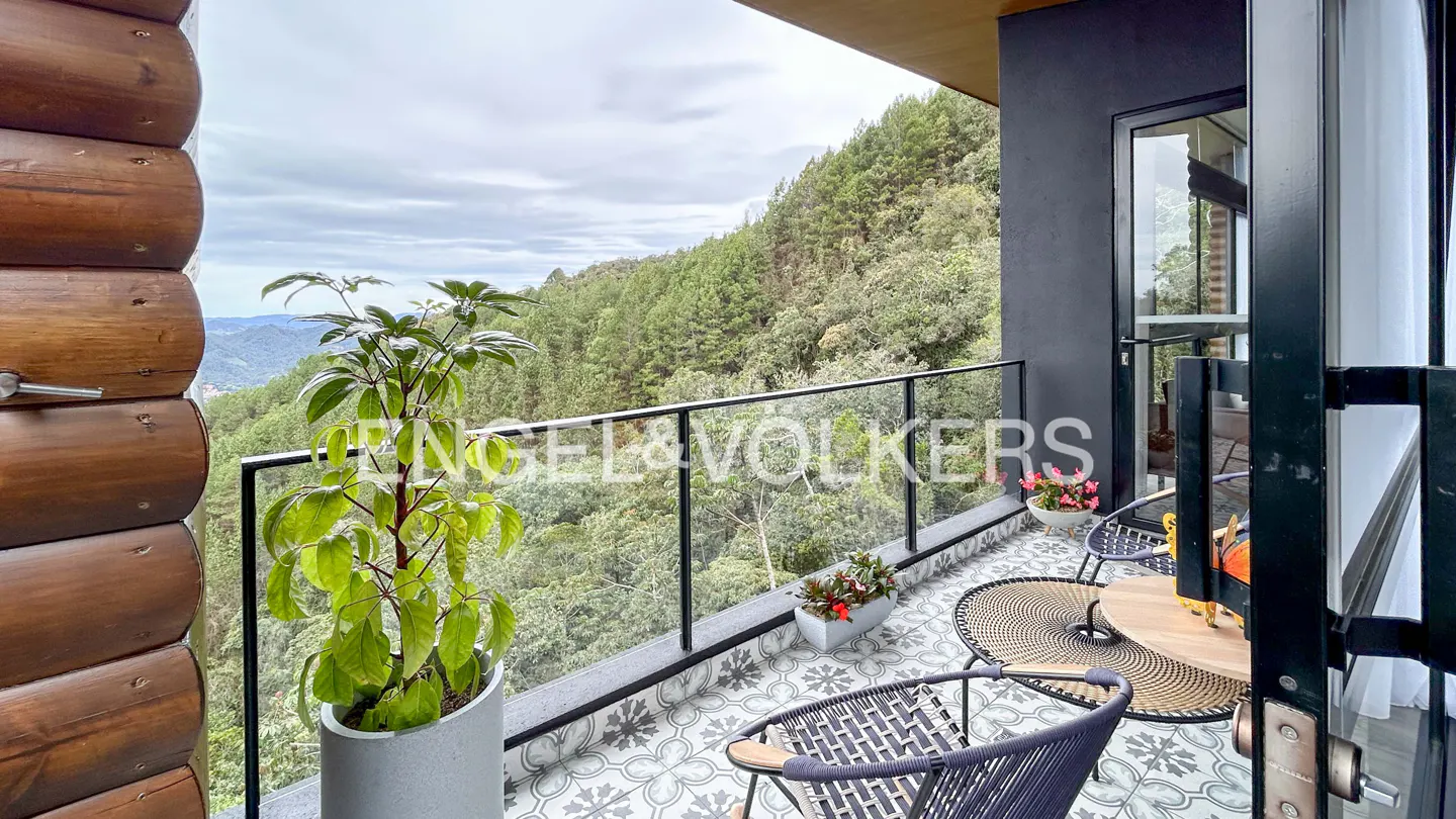 Balcony view with chairs, table, and plants overlooking a lush green forest. The balcony has a glass railing and patterned tile flooring.
