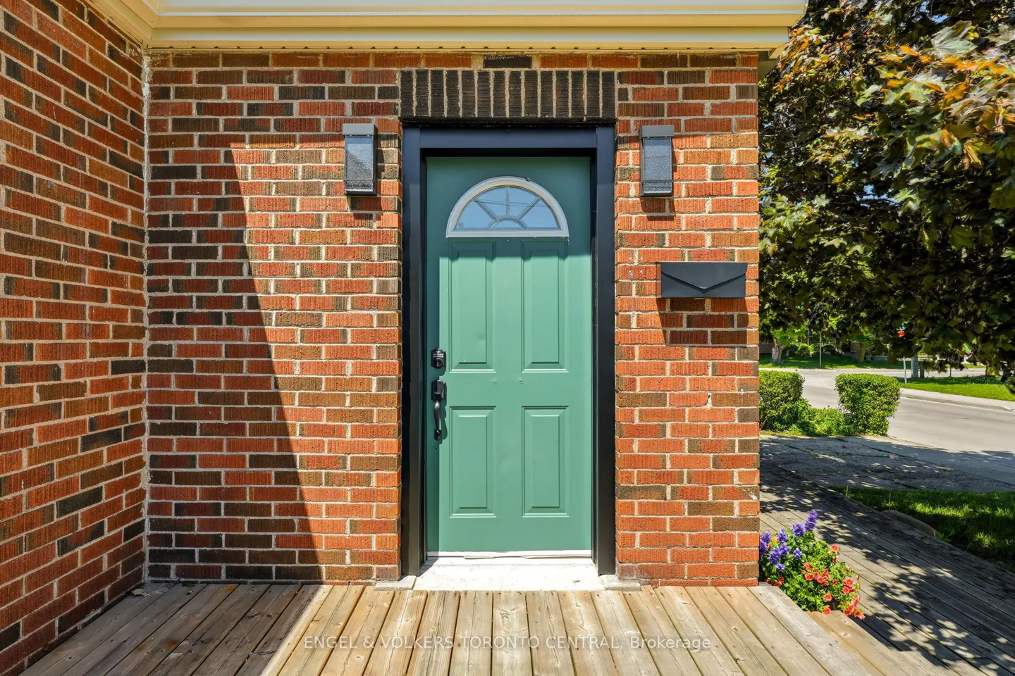 A sage green front door with a semi-circular window, set in a red brick wall with a wooden porch and black mailbox.