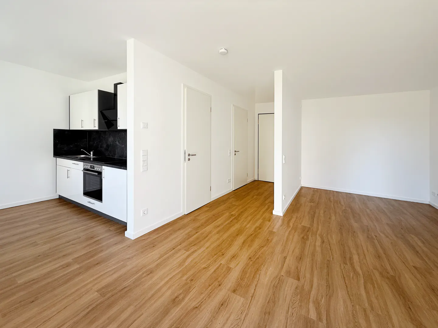 Bright, empty apartment with wood floors, white walls, and a modern kitchen with black countertops. Three white doors are visible.