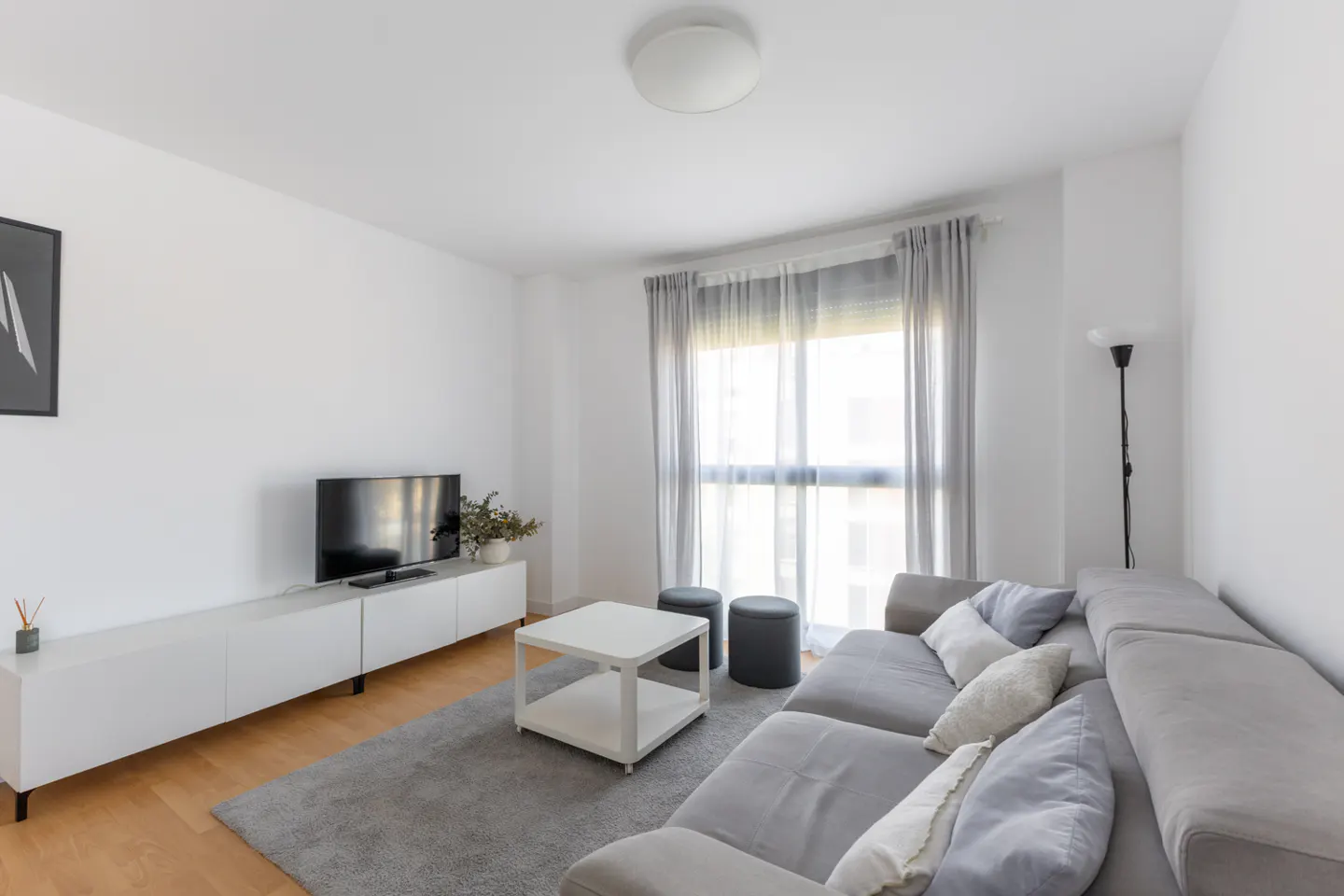 Bright living room with gray sectional sofa, white walls, and wood floors. A TV sits on a white console table. Gray rug and curtains.